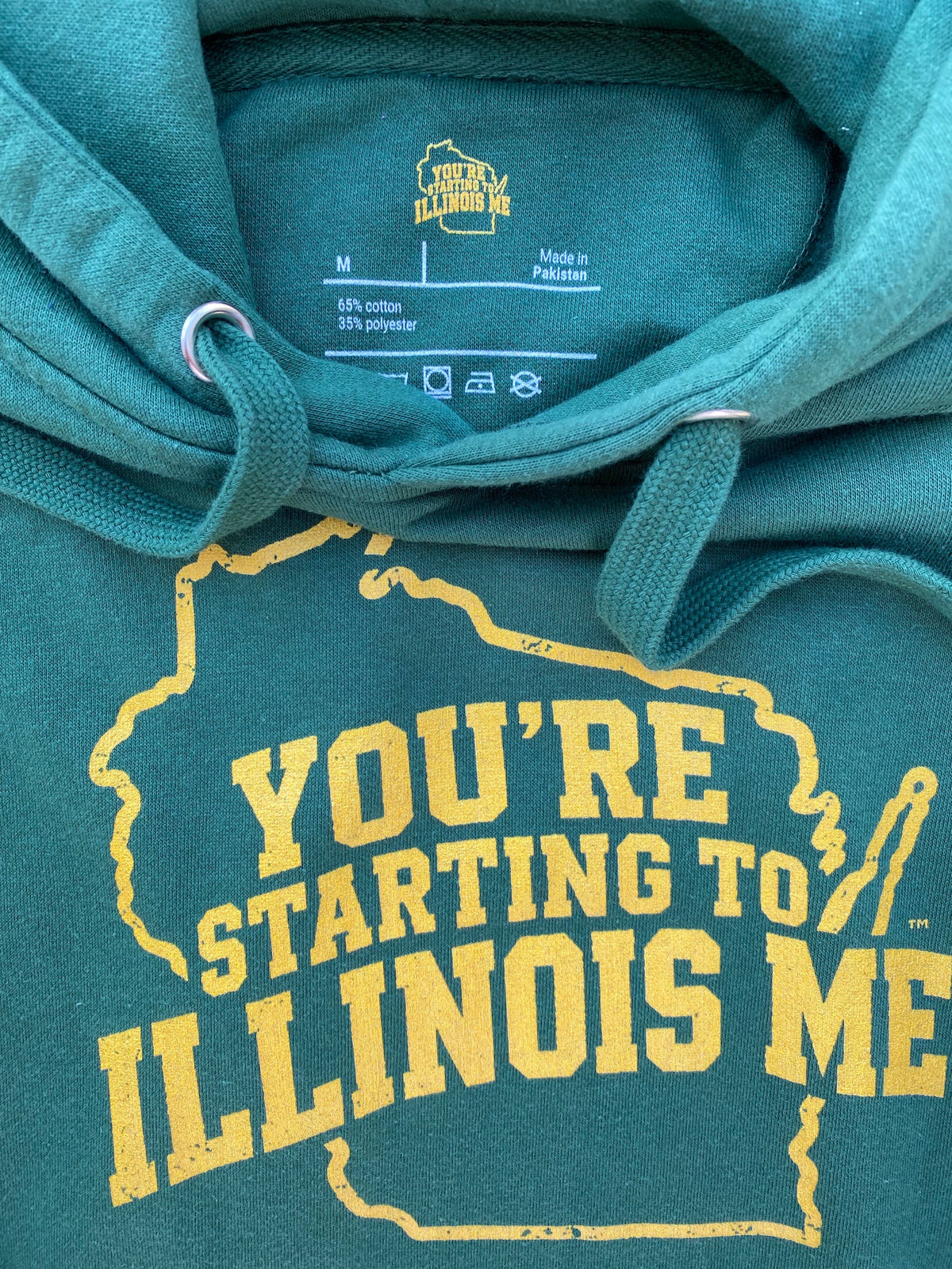 Close-up of a green hoodie with gold “You’re Starting to Illinois Me” lettering, a Wisconsin outline, and branded neck print.